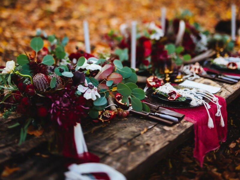 An outdoor table at a fall wedding complete with beautiful flowers in shades of red, white candles, and black dinner plates.