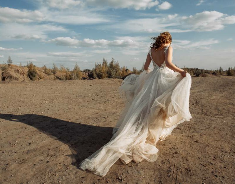 A bride with curly brunette hair stands in the desert with her back turned to the camera. The wind billows her dress.