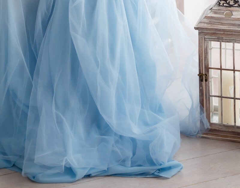 The hem of a blue puffy wedding dress inside a room during the day. Next to the dress are two unlit lanterns.