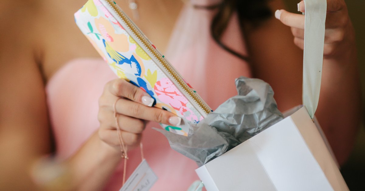 A bridesmaid takes a pink paisley print wristlet from a gift bag. A small white tag is attached to a pink string in her hand.