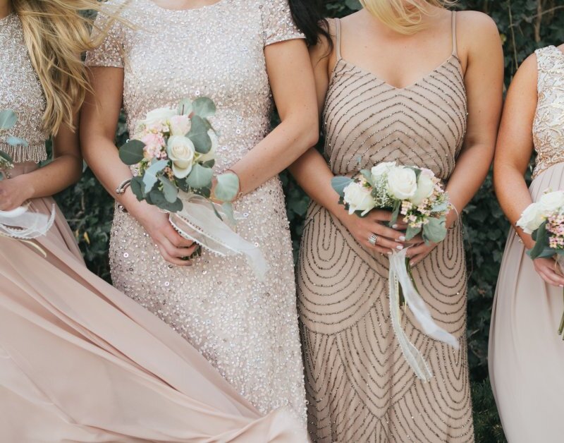 Four bridesmaids wearing different dress styles stand in a line against a bush. All four hold bouquets in their hands.