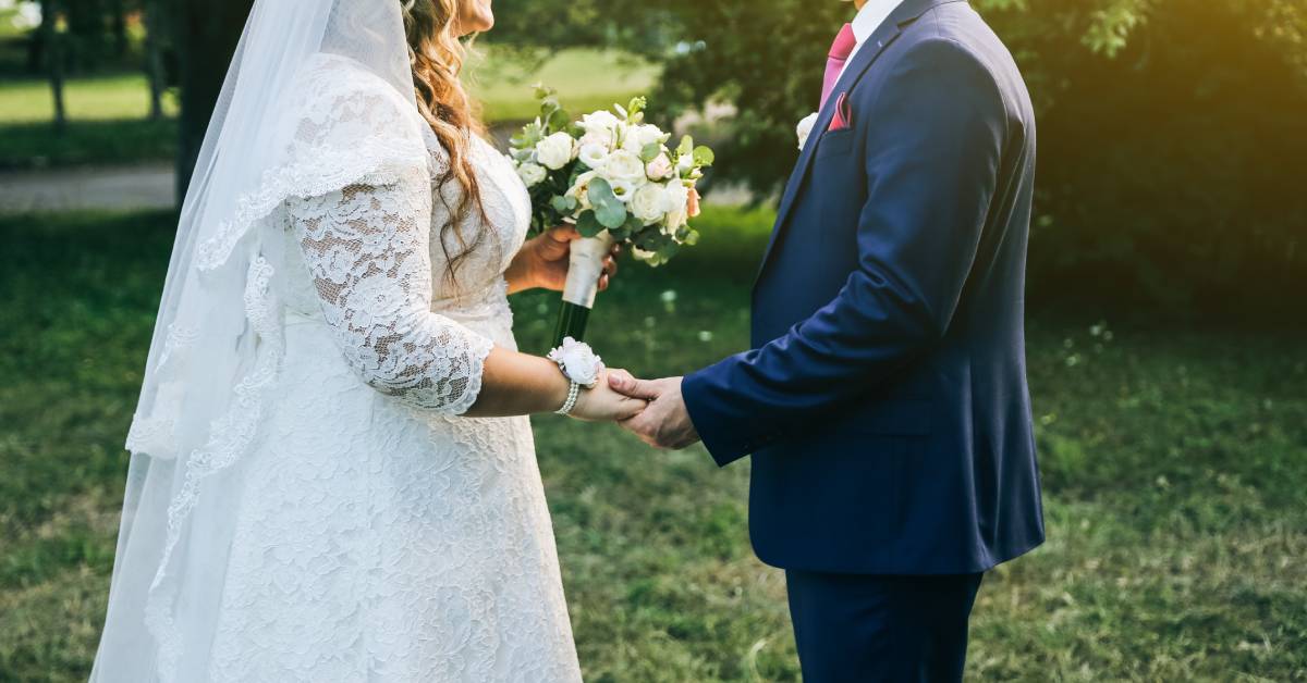 A bride and groom stand in a park holding hands. The bride holds a bouquet in the other hand; the background is grassy.