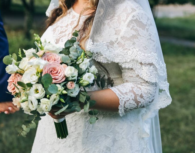 A bride wearing an hourglass dress has her arm intertwined with another person. She has a bouquet in her hand.