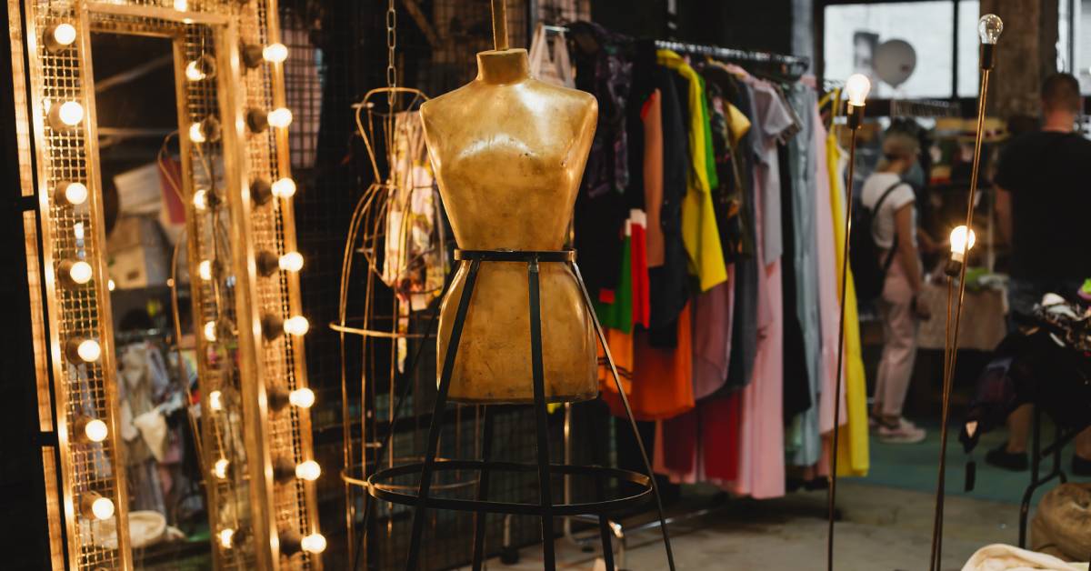 A mannequin is center on display at a boutique shop. There is a large mirror with bulbs and a clothing rack in the background.