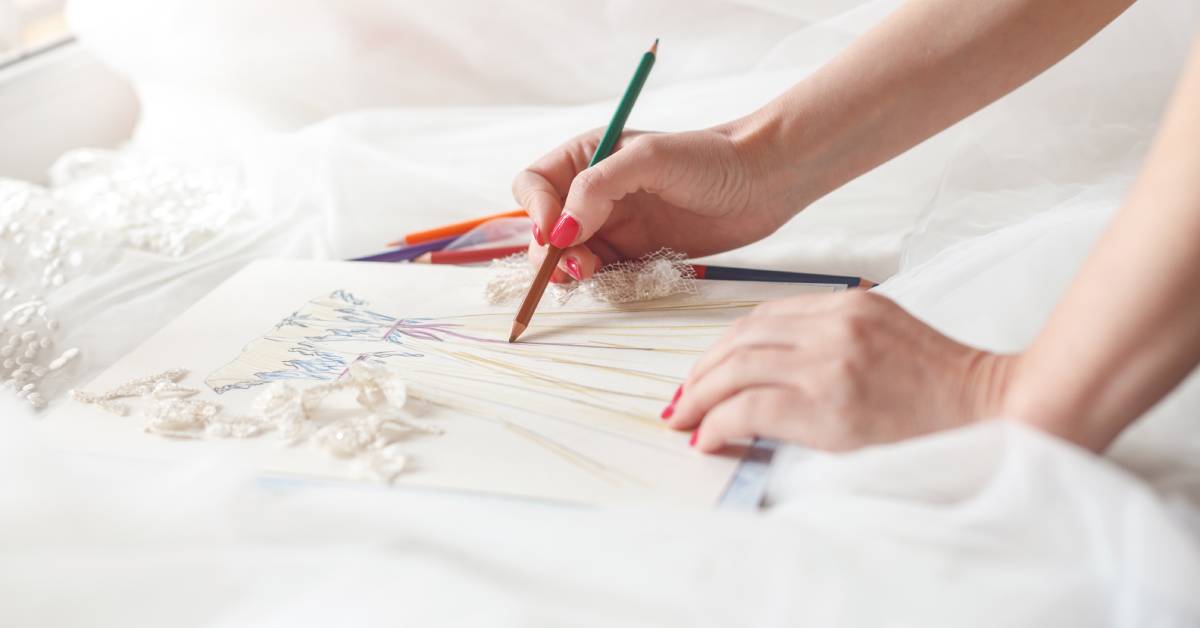 A dress designer sketches out her ideas on a paper and adds details. There are fabrics and materials around her.