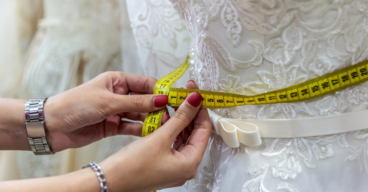 A close-up shot of hands taking measurements around the waist of a white wedding dress. There are floral patterns on the gown.