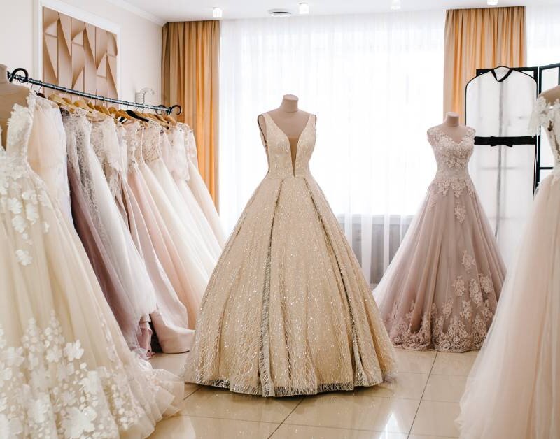 Four mannequins decorated with elegant dresses are spread out in a fitting room. Bright sunlight pours in from the window.