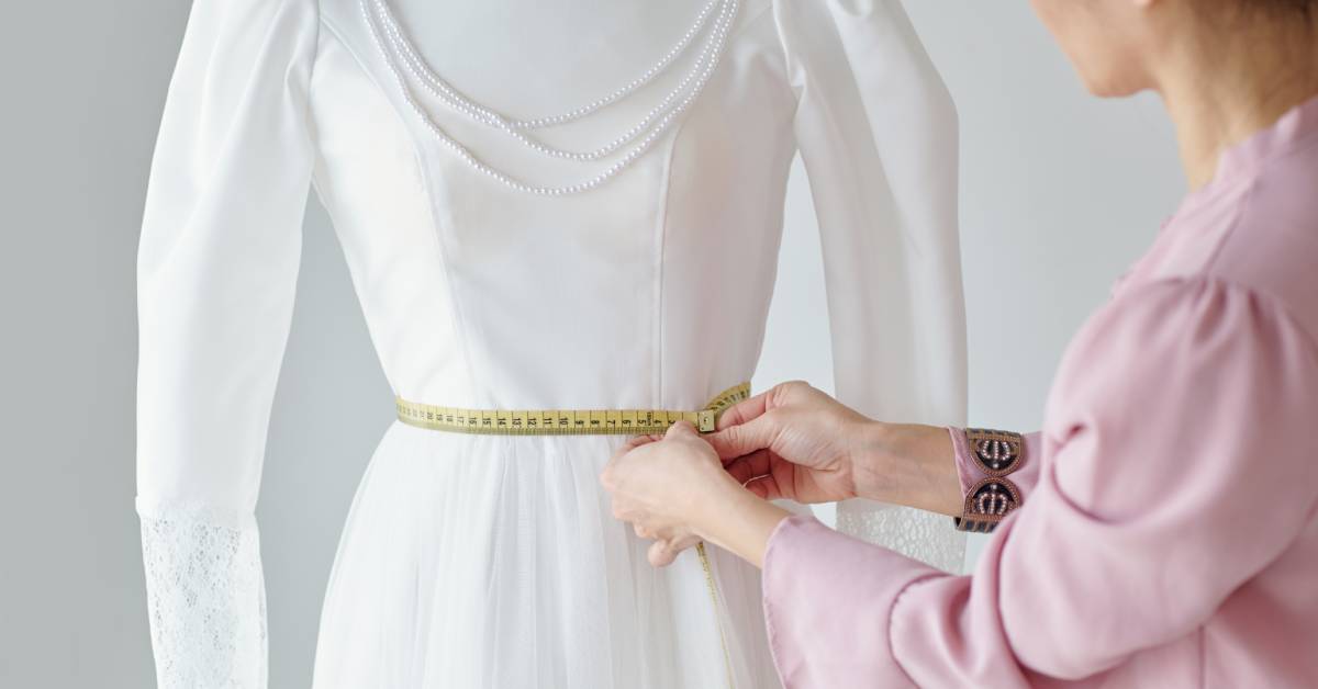 A seamstress wearing a pink shirt measures the waistline of a white wedding dress on a mannequin with pearls.