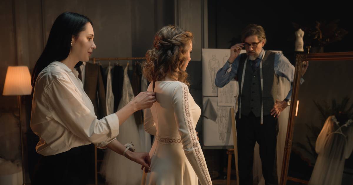 A woman stands in front of a mirror as a tailor zips up her dress from behind. Another tailor supports from a distance.