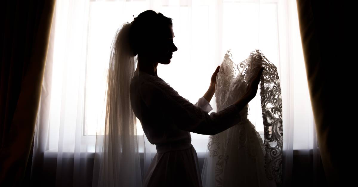 A silhouette of a bride against a large window, preparing to dress up in her wedding gown as she holds it in her hands.