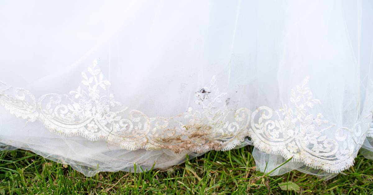 The bottom of a white wedding dress is stained with dirt and smudged on a small area as the bride stands on grass.