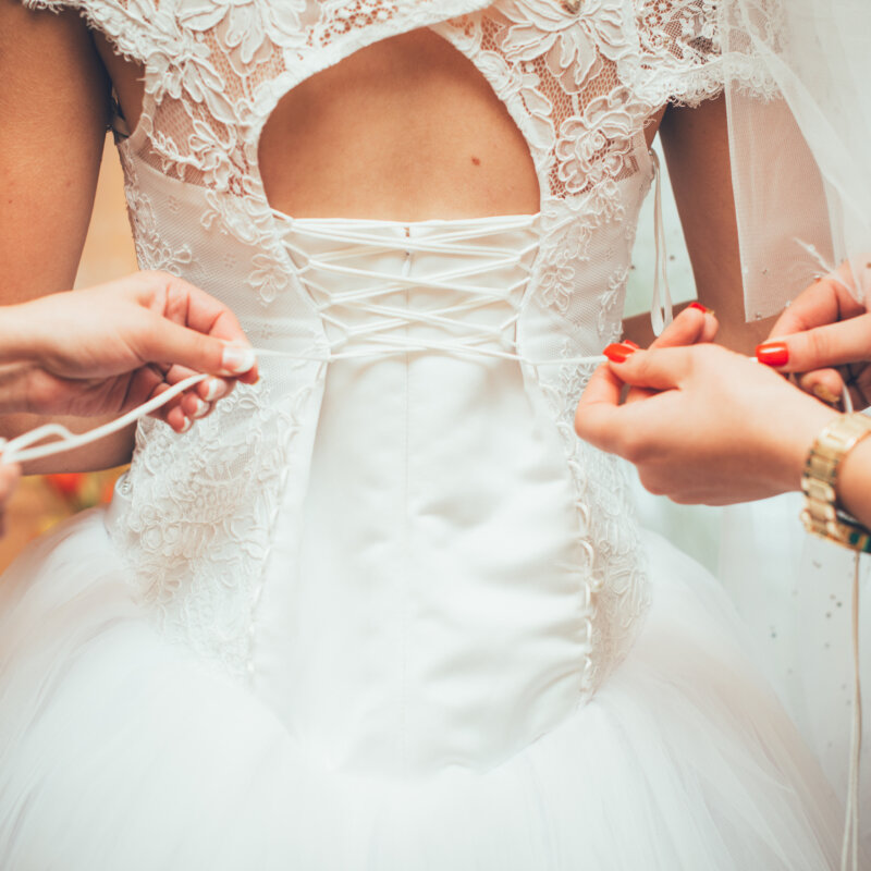 A bride standing in place as two assistants finalize her final dressing, tying the lacing on her back.