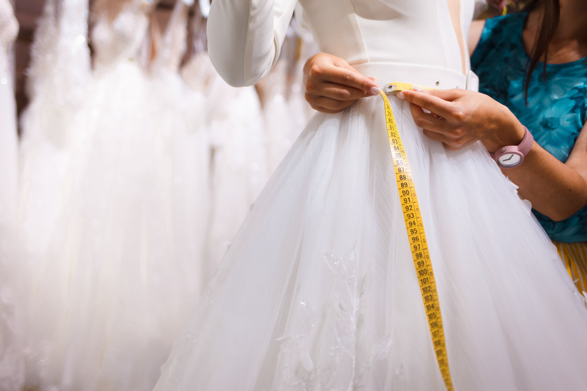 A professional tailor wrapping a flexible measuring tape around the bride's waist. Several dresses hang in the background.