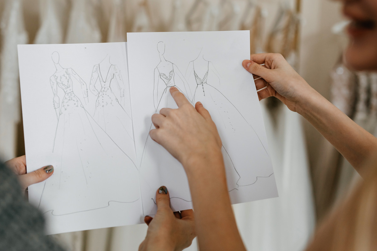 A designer holding up a sketch of a wedding dress as she faces the bridal client and they discuss details of the process.