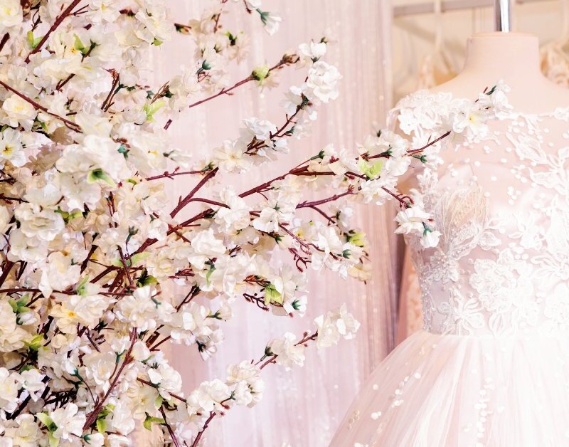 An elegant gown is exhibited on a mannequin next to a decorative plant. There is a curtain in the background.