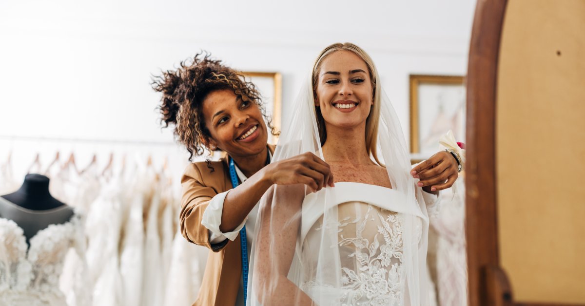 A tailor smiles as she helps her bridal client with her fitting. They are in front of a mirror in a boutique shop.