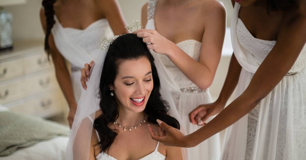 Three bridesmaids in a bedroom with the bride, helping her get ready. They place delicate flowers on her veil.