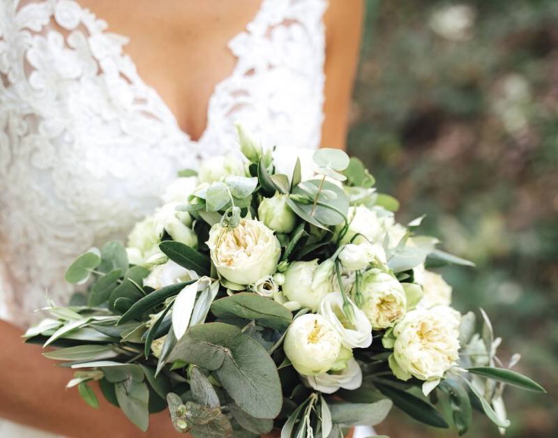 A bride holding a white and green bouquet in her hands against her white wedding gown. Her ring is visible on her finger.