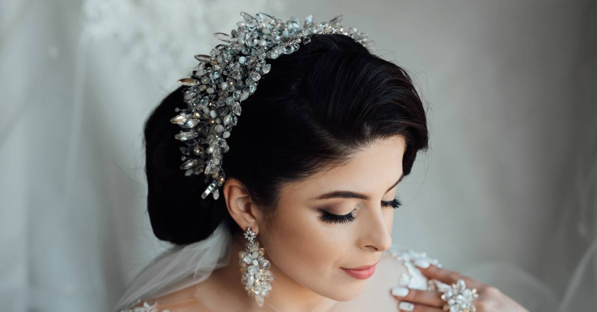 A clear crystal crown on top of a bride's hairdo. She wears matching crystal earrings and a matching statement ring.